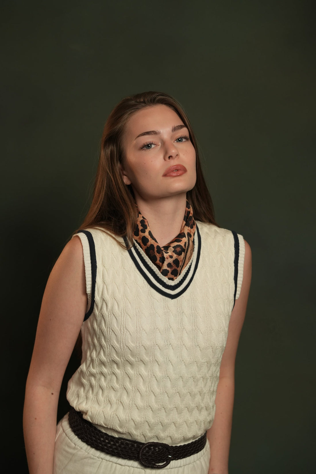 Woman in a cream cable-knit sleeveless sweater with a navy-striped V-neck, a leopard-print scarf at the neck and a woven belt