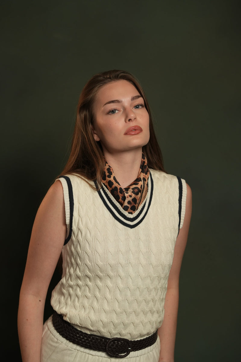 Woman in a cream cable-knit sleeveless sweater with a navy-striped V-neck, a leopard-print scarf at the neck and a woven belt