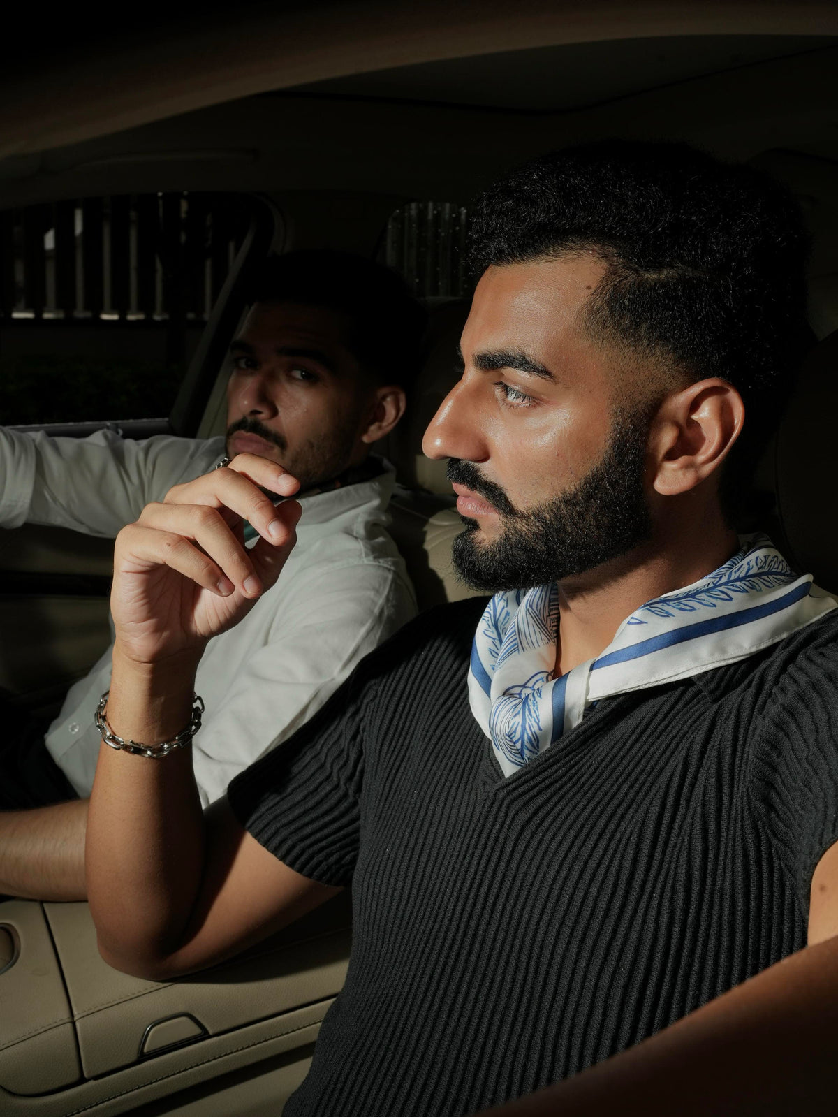 Two men sitting inside a car, one with a blue scarf around his neck.