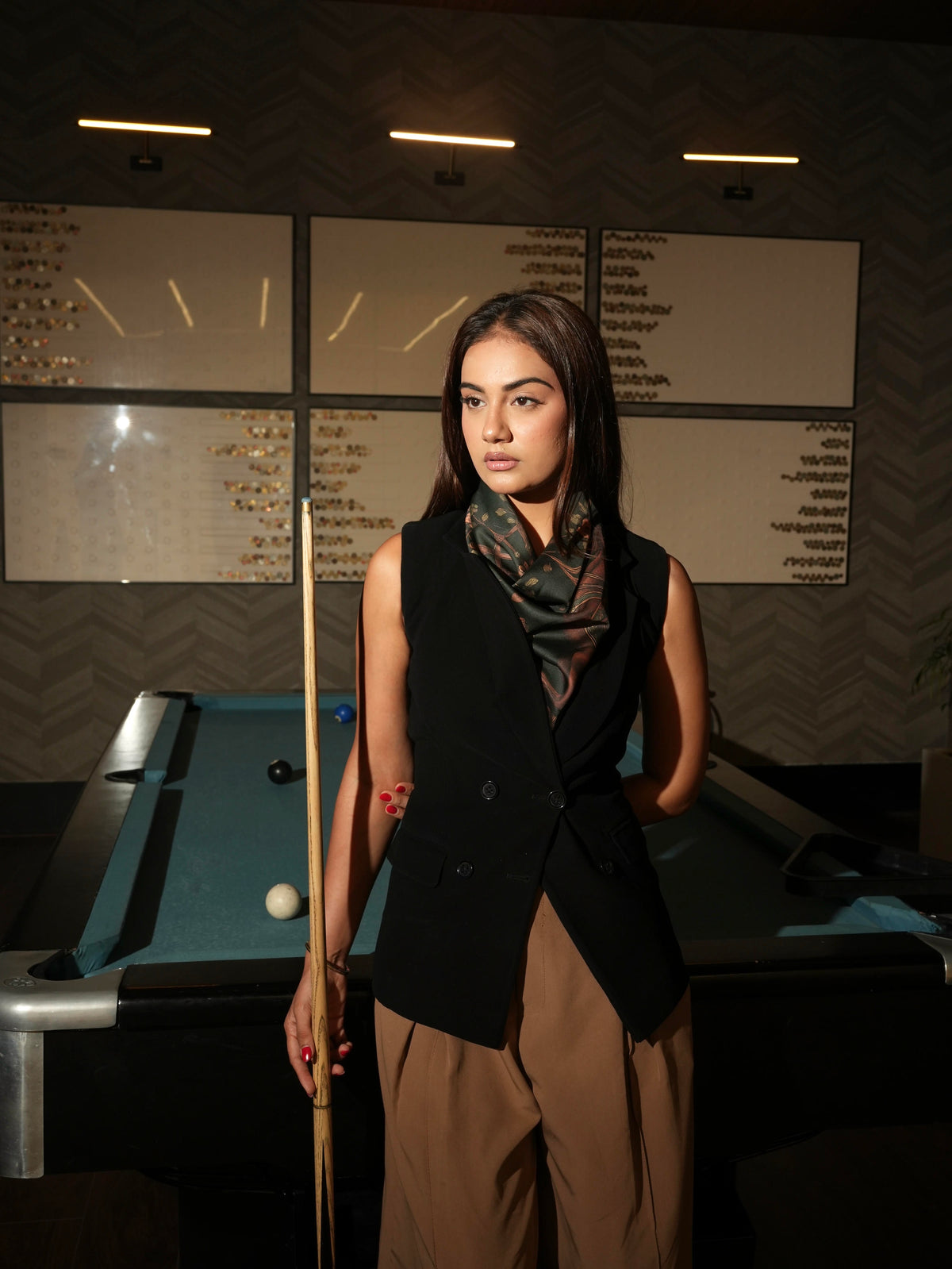 Woman wearing a dark pattern scarf in front of a pool table in a dimly lit room.