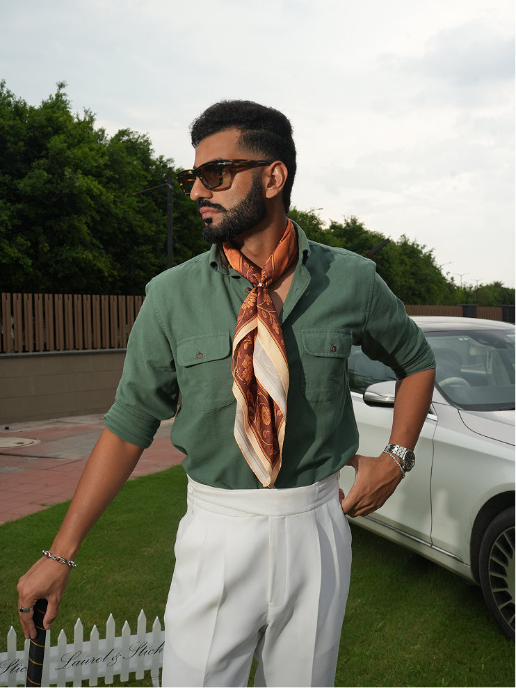 Man wearing a green shirt, white pants, and an orange scarf outdoors with a car in the background.