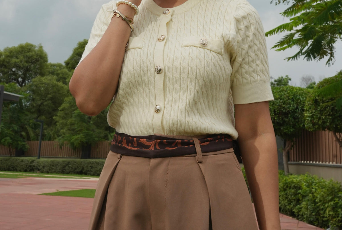 Person wearing a cream knitted cardigan and beige skirt with a scarf as belt in an outdoor setting.